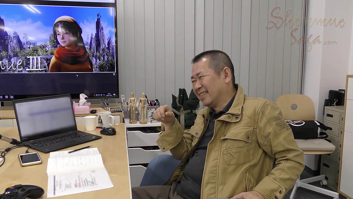 Suzuki lächelnd am Arbeitsplatz mit Notebook, Notizen und Holzfiguren im Hintergrund