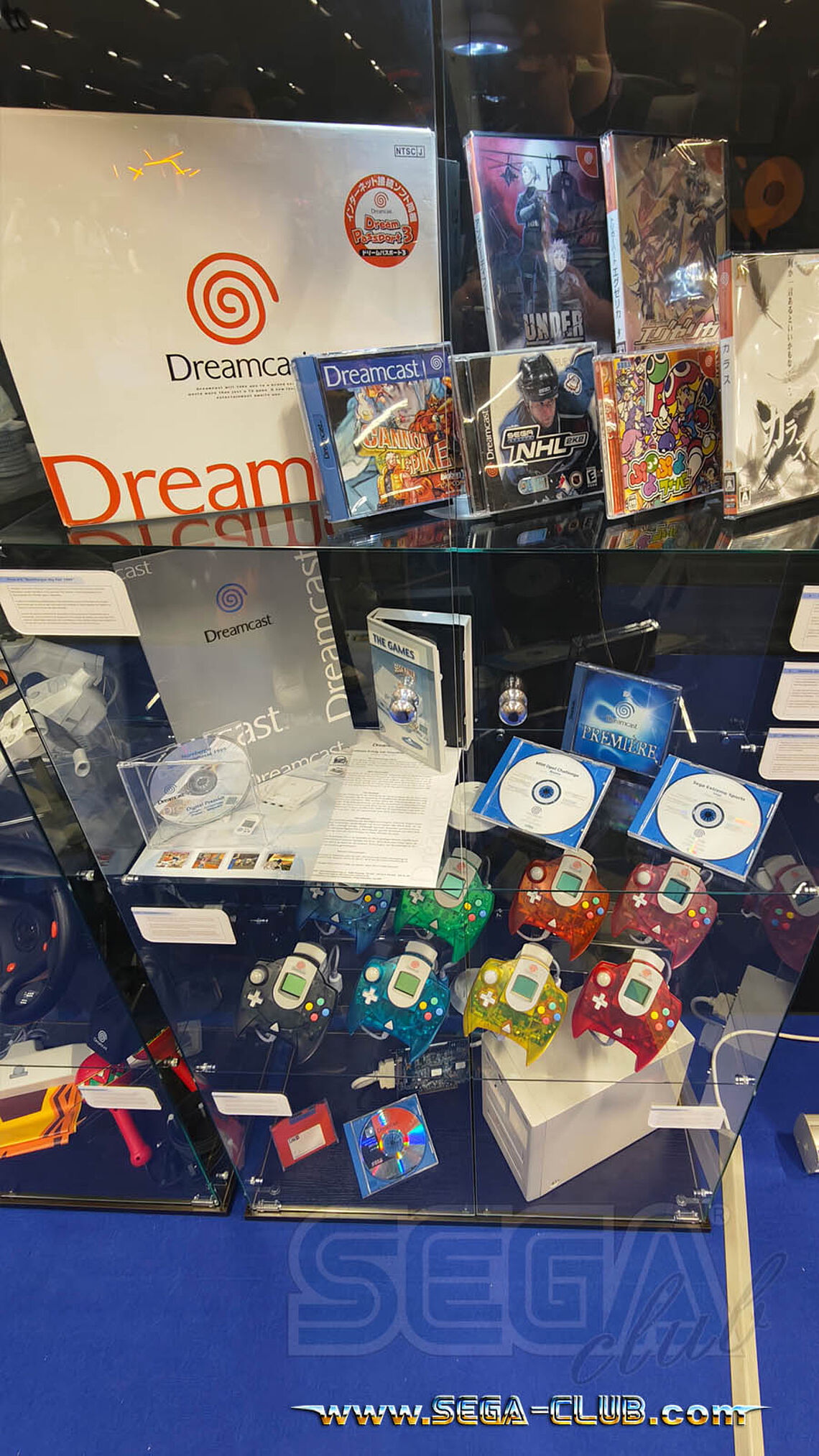 Vitrine mit Spielen, Kartons und Hardware: acht farbige Dreamcast-Controller, Maracas-Rasseln und mehr