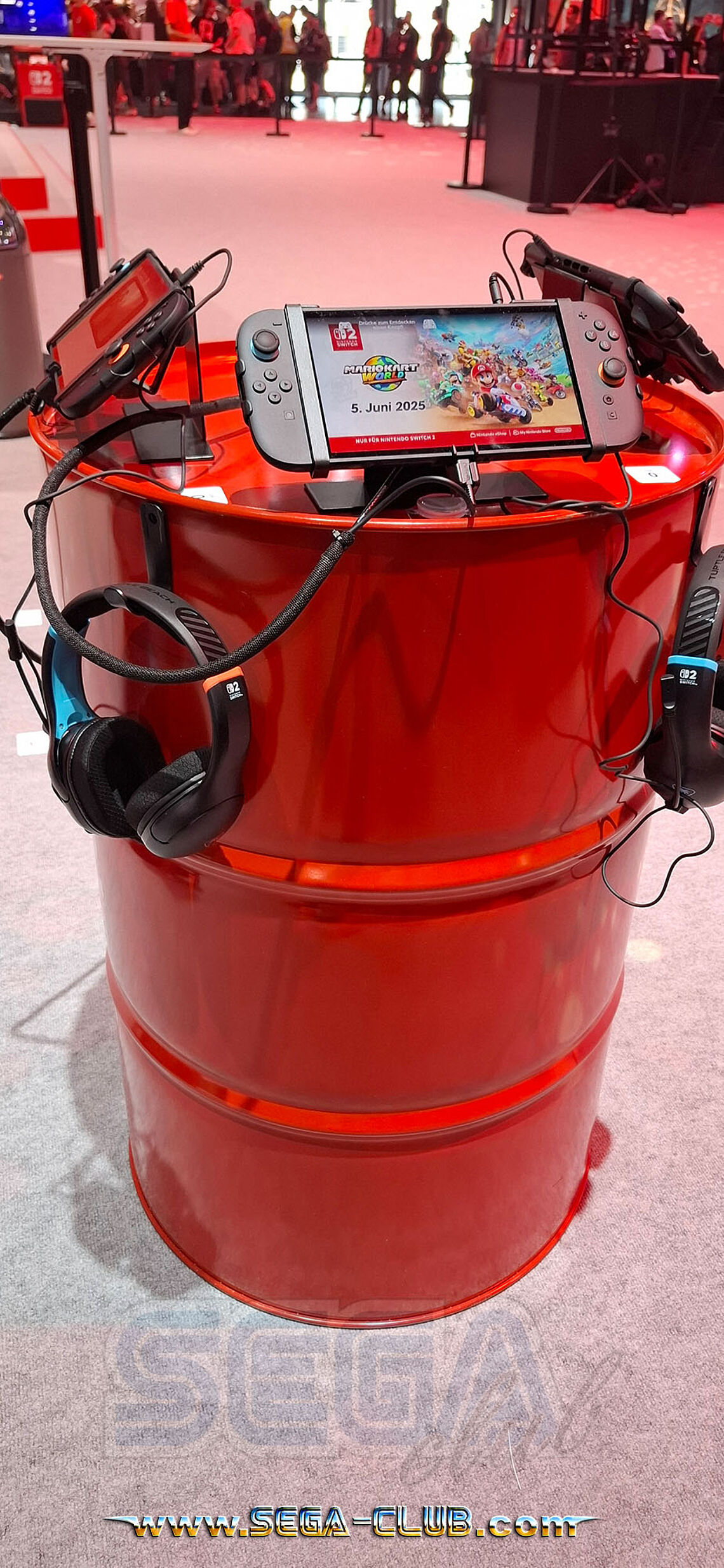 Rote Tonnen am Nintendo-Stand beherbergen angeschlossene Switch 2 Systeme und Kopfhörer mit Mario Kart World