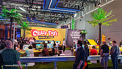 Blick über die Fahrbahn-Leitplanke auf den Crazy Taxi World Tour Spielbereich umrahmt von grünen Palmen und gelben Tischen